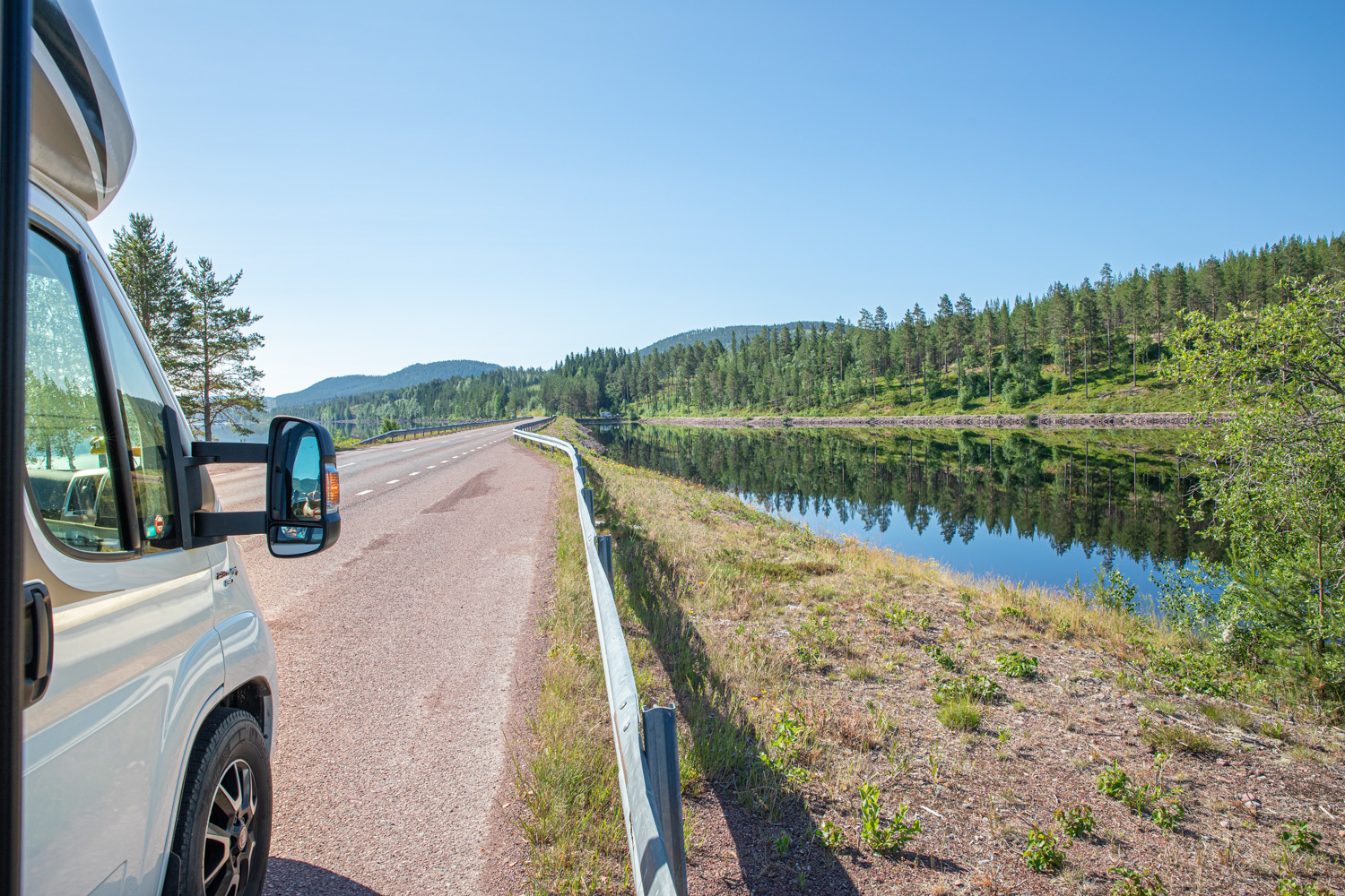 Fronten på en husbil ses på en väg med en bro över vatten. Bortom vägen syns skog och berg i bakgrunden.