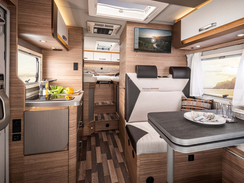 Interiört i en husbil från Weinsberg. En sittgrupp vid ett fönster. Här syns ett bord samt tvåsitssoffa med rak rygg mot vägg till höger i bild. Till vänster, där ingången är, syns en köksmodul. Längst bort i bild ser man två höga sängar.