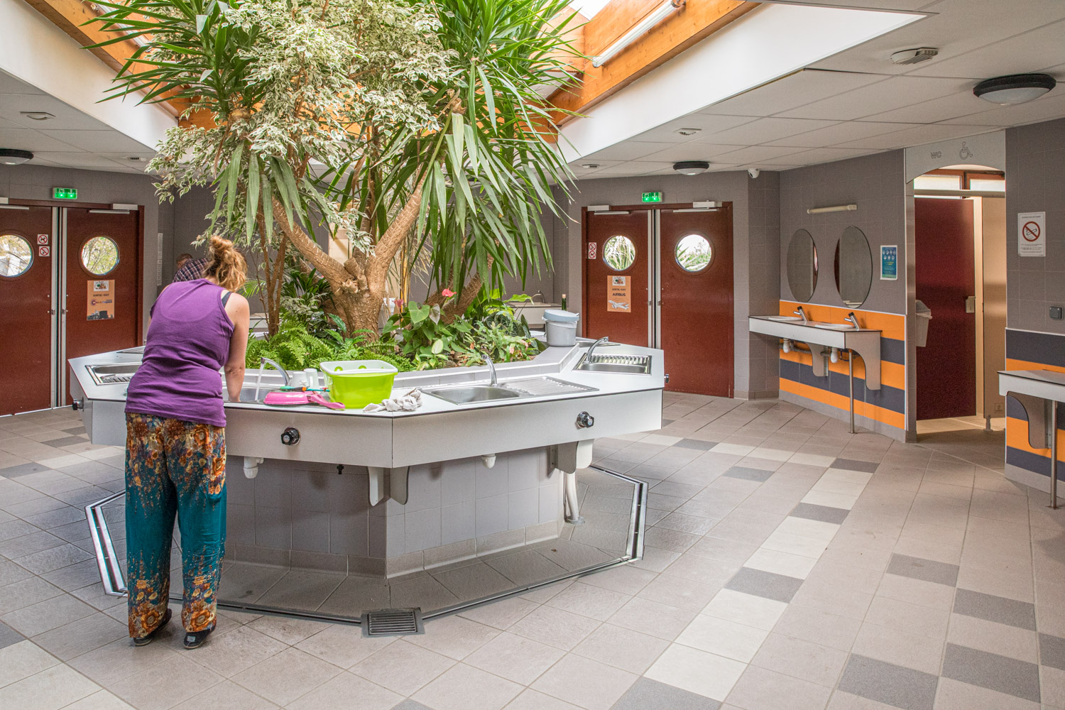 Servicehus på camping med toaletter på sidorna och en åttakantig köksö med diskhoar och ett trä i mitten.