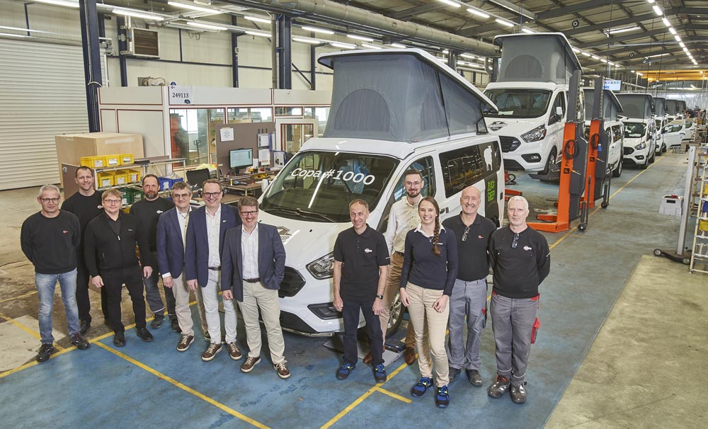 Husbilsfabrik med minst sju husbilar på rad på produktionslinjen. Den första har Copa #1000 skrivet på rutan. Framför bilen står olika chefer och ser glada ut.