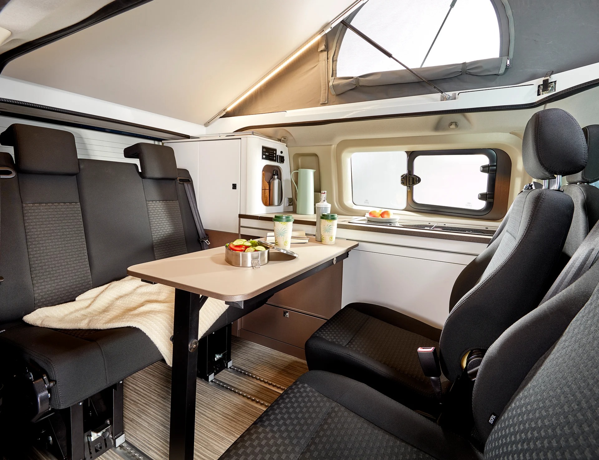 Kompakt husbil med två enkelsäten och en soffa vända mot varandra. Man anar även en förarstol bakom en av passagerarstolarna. Liten köksmodul syns.