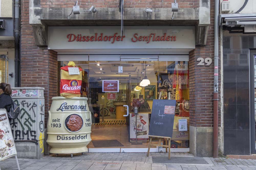 Butiksfasade med skyltning i form av stora senapsburkar utanför. Samt en skylt om att bara fyra kunder är tillåtna i butiken samtidigt.