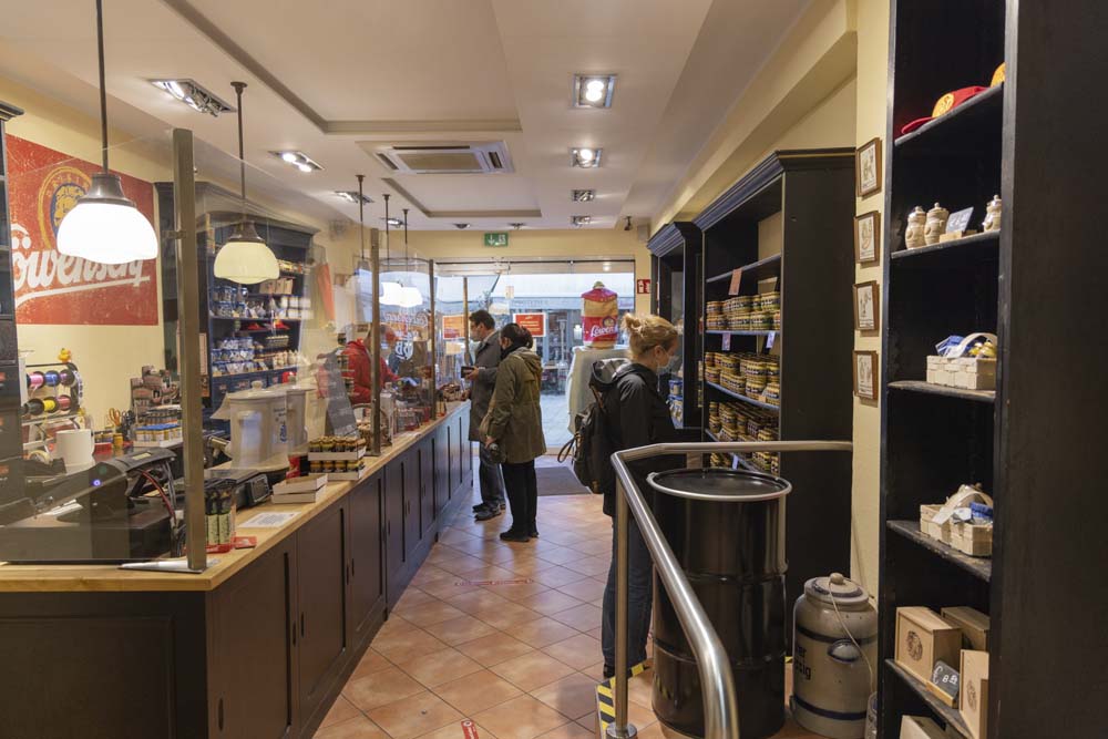 Butik med en lång disk med glasruta framför syns till vänster i bild. Till höger finns hyllor i trä med allehanda varor. Tre kunder ses i butiken.