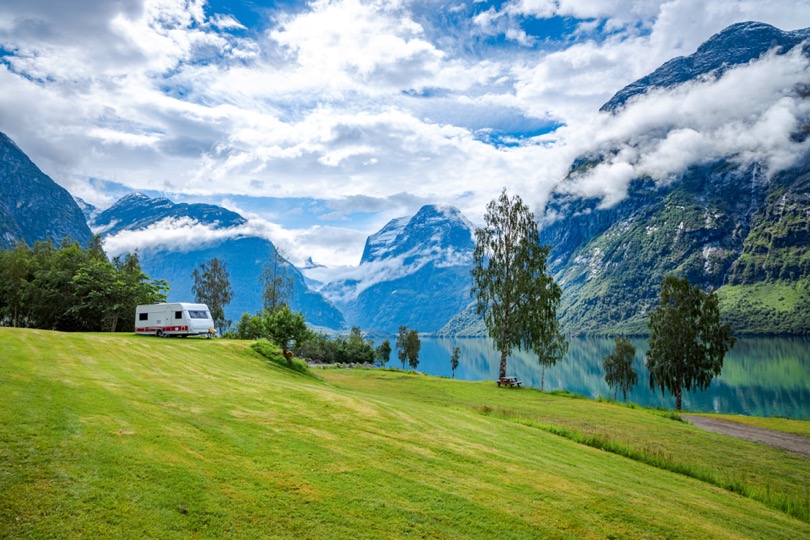 En husvagn står på en gräsmatta med några träd och berg bakom sig. Till höger i bild anas en väg och en sjö.