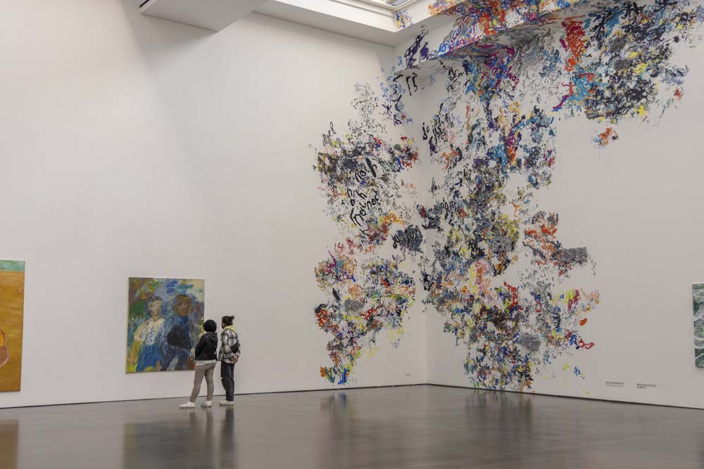 Stort vitt rum på museum med väggkonst i ett hörn. Målningen letar sig upp mot taket där det finns ett takfönster.  Bredvid står två människor och tittar på en tavla.