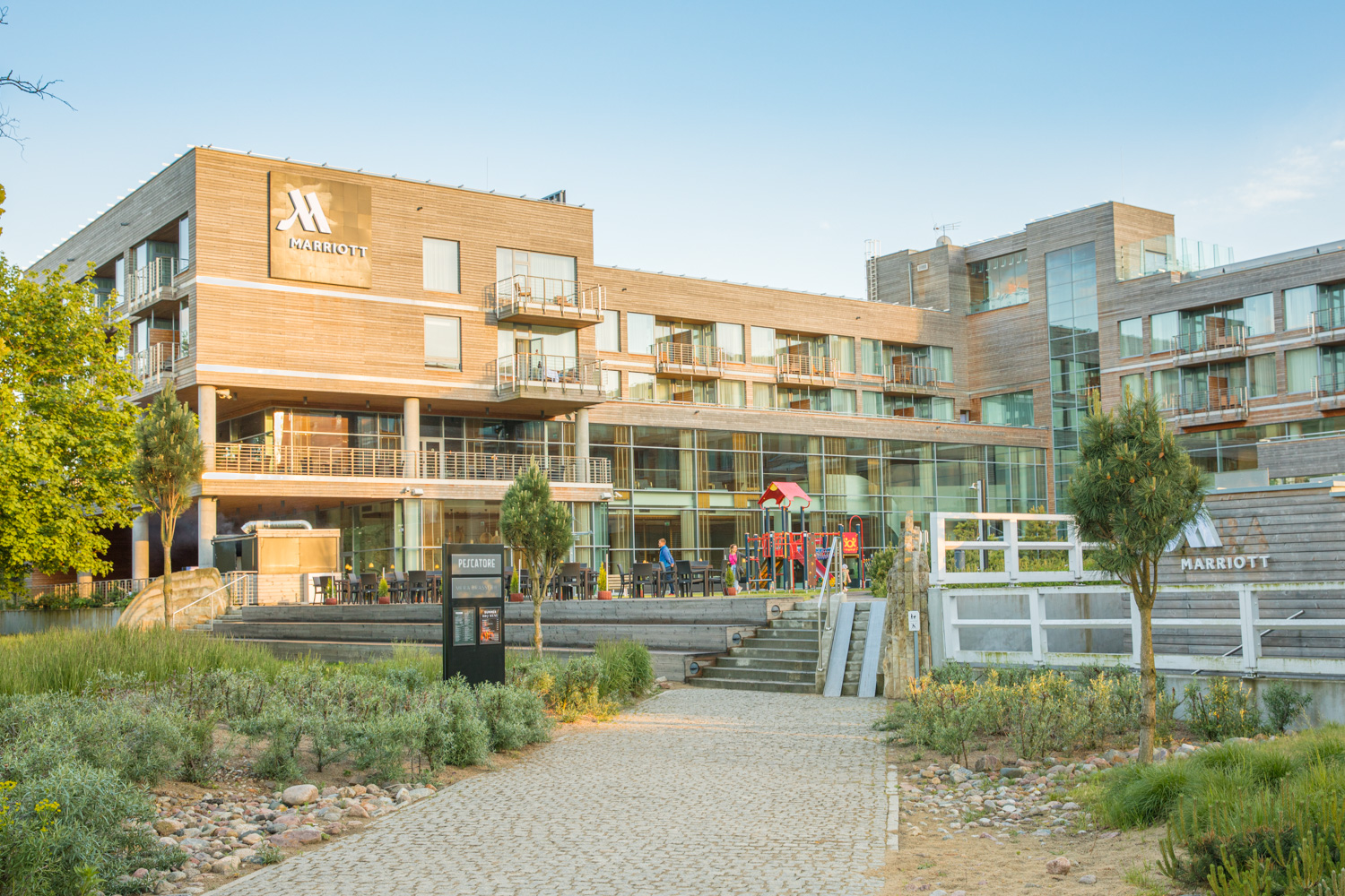 Hotell i trä och stora glaspartier. Framför hotellet finns en lekplats och en uteplats med bord.