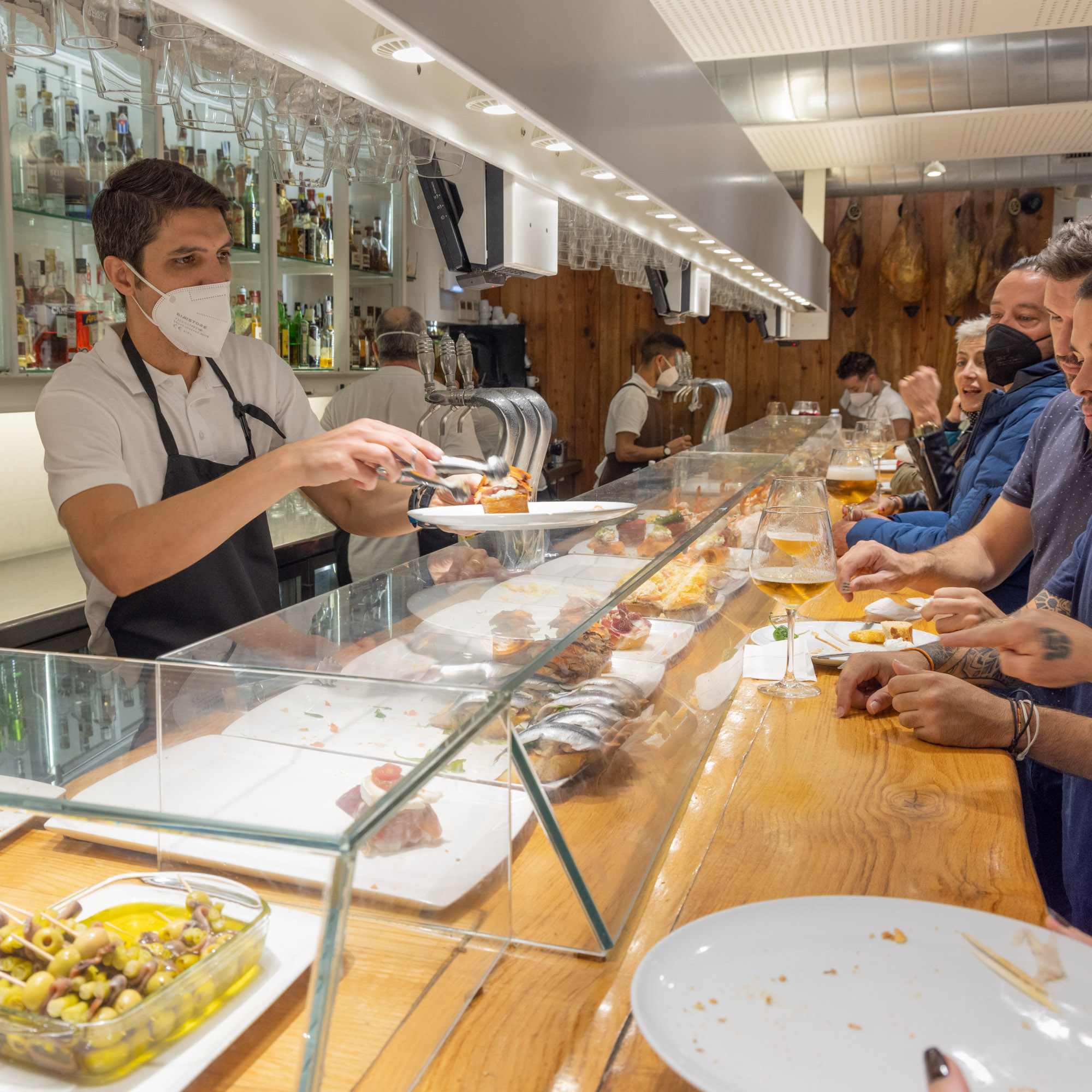 Ljus bar med tre män i vita pikétröjor med svarta förkläden står bakom baren och expedierar de många kunderna som står rätt nära varandra på andra sidan baren.