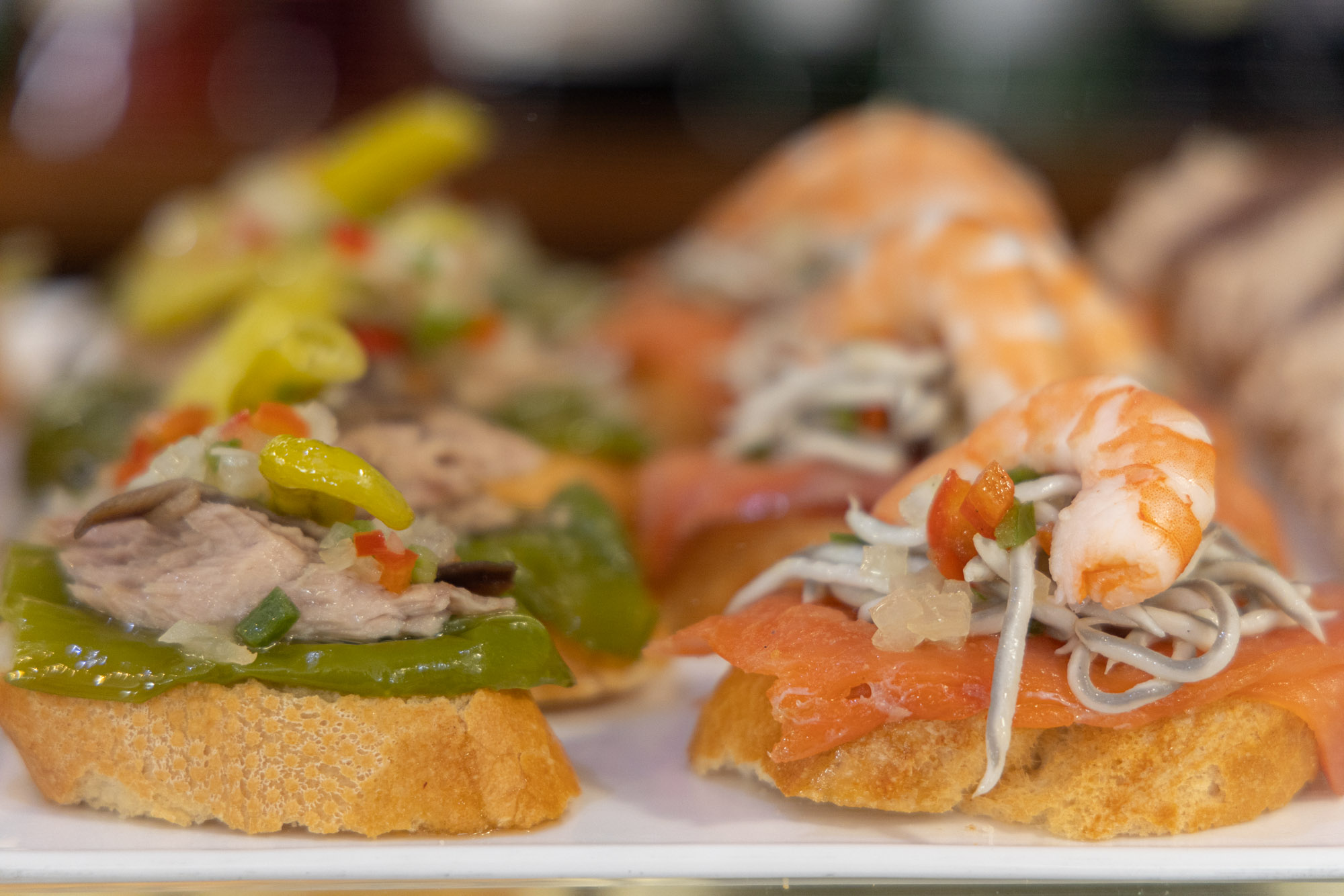 Flera tapas på en tallrik med två stycken i fokus. Båda har baguette som bas och toppas med fisk och skaldjur.