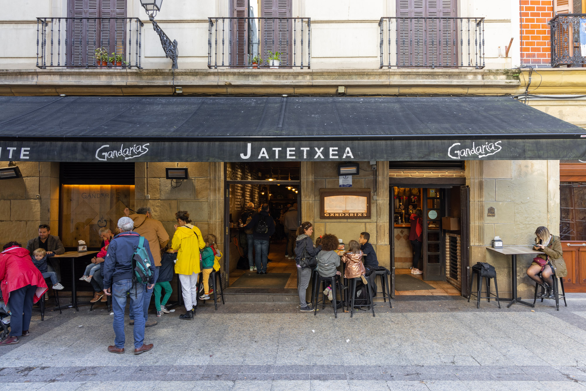 Restaurang Jatetxea i San Sebastián. Bild sedd från gatan. Utanför ses fyra bord. Det sitter någon eller några vid varje bord. Man anar baren innanför de två pardörrarna till höger. I dörren till vänster ser man att några står och väntar på sin tur att få ett bord.