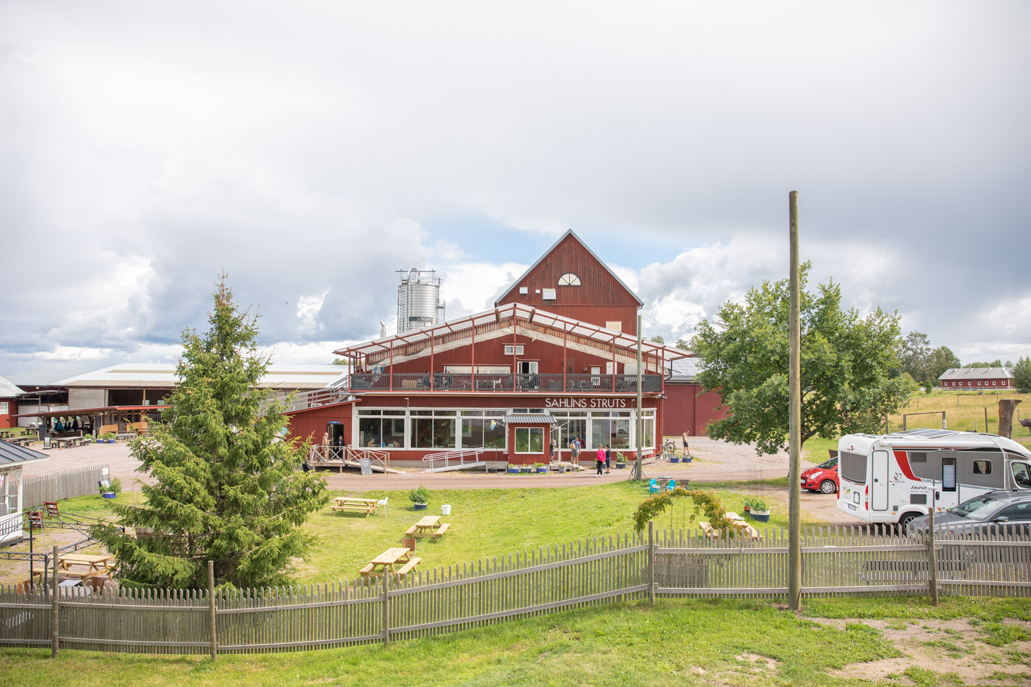 Röd byggnad i trä med vita fönster och en takterrass. Bakom syns ett ladutak. Framför huset finns en gräsmatta med picknickbord och till höger en stor parkering.