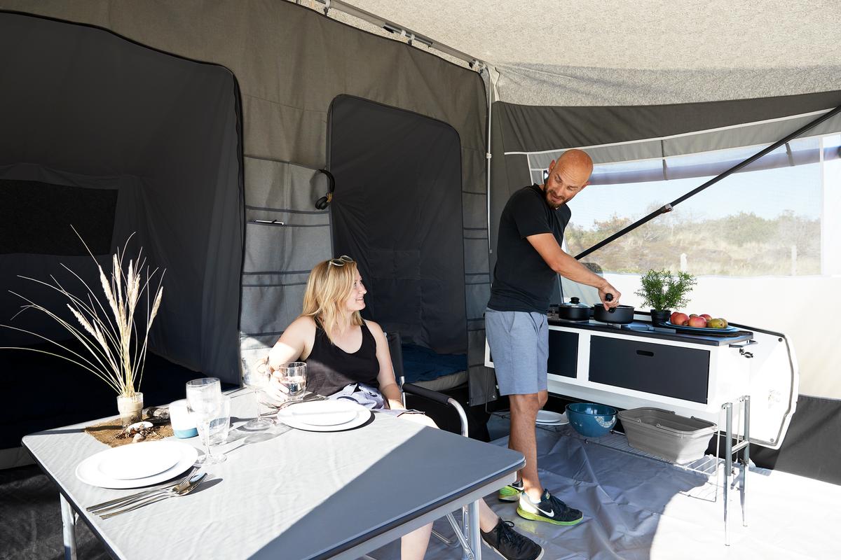 En man står och lagar mat i Camp-let North medan en kvinna sitter och väntar vid ett dukat bord. Bakom henne syns två sovalkover.