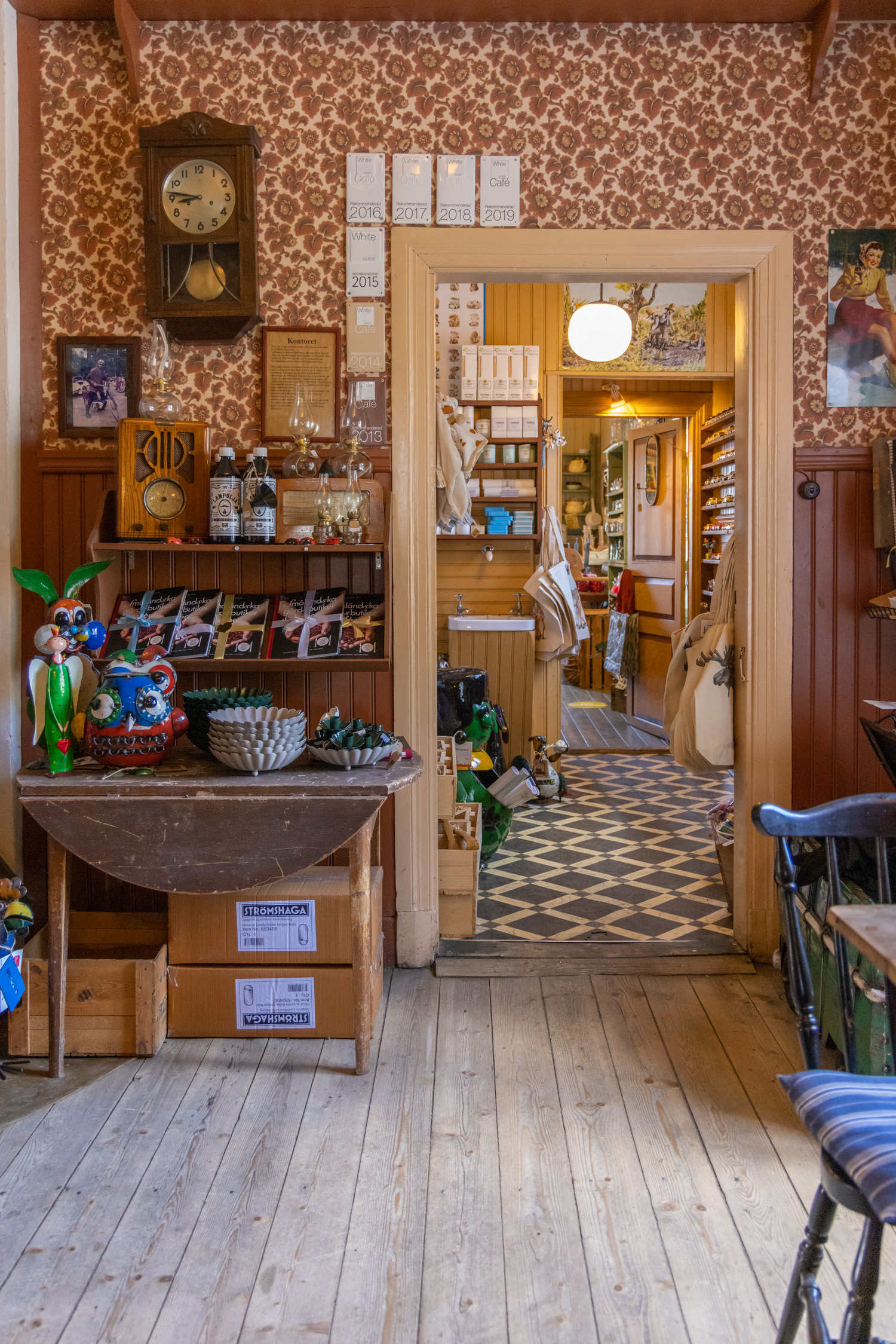 Från ett rum ser man en hall och ytterligare ett rum. Alla rummen har olika golf och väggbeklädnad. På väggen i första rummet finns en brunblommig tapet och ett bord med varor på.