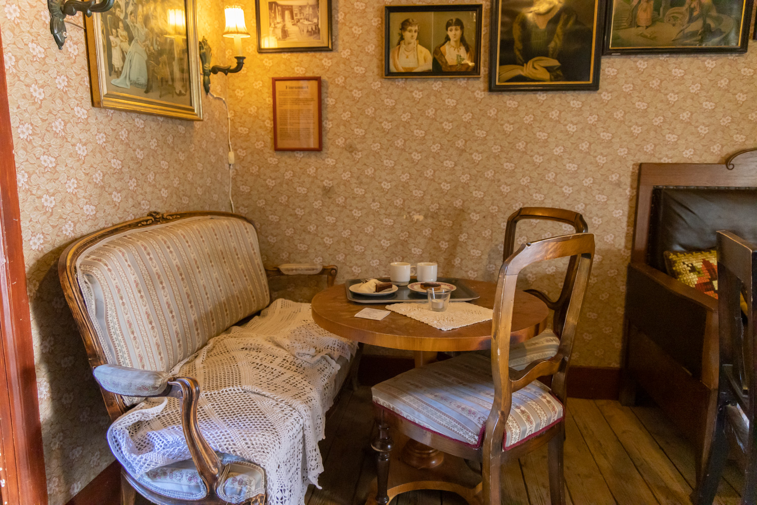 Ett mysigt hörn med en gammal soffa, ett runt bord och två stolar. Ännu en soffa och en stol skymtas i utkanten av bilden. På väggarna finns gamla fotografier och lampor. Tapeten är blommig i beige.