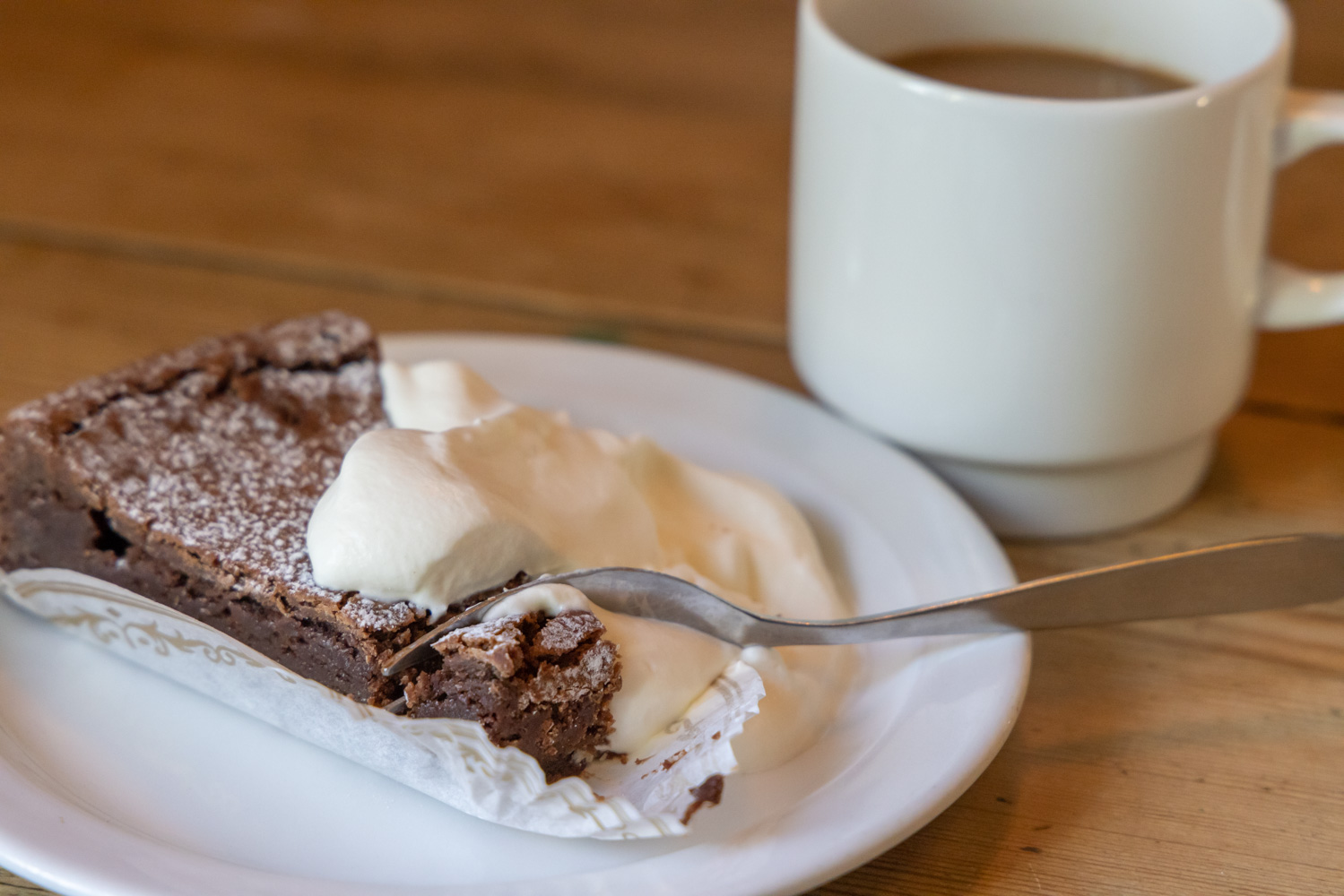 En krämig kladdkaka ligger på ett fat med vispad grädde. Bredvid finns en kopp kaffe med mjölk.