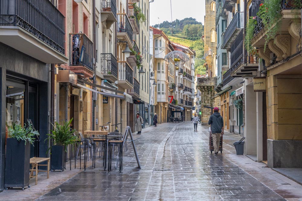 Smal gata med höga äldre hus med vackra balkonger. På gatan syns bord och stolar utanför en restaurang.